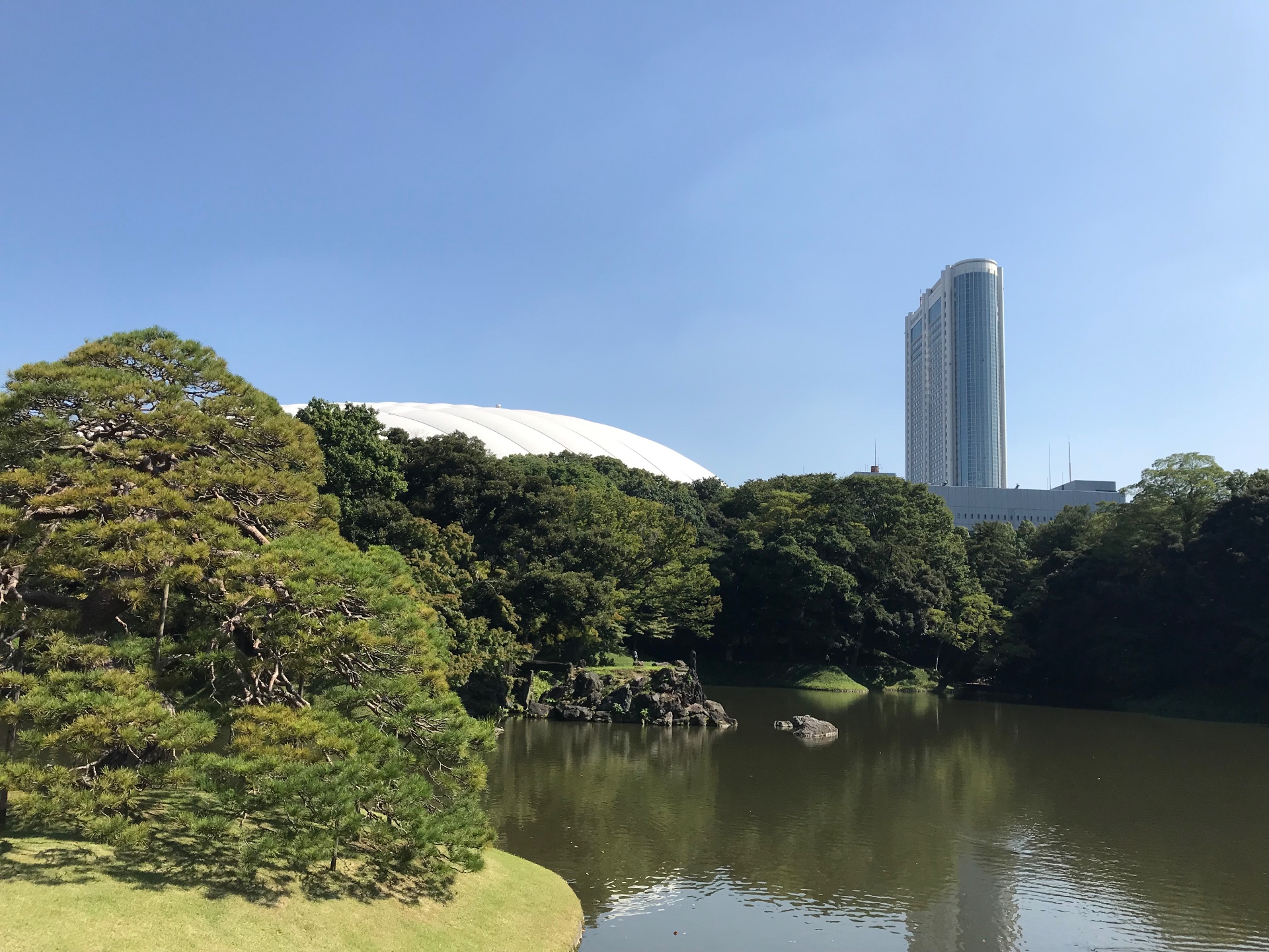 Teich im Koishikawa-Kōrakuen-Garten mit Kiefer im Vordergrund und Tokyo Dome im Hintergrund